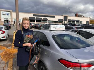 A smiling young woman holding a stuffed animal stands beside a car in the parking lot of the Best Drivers Academy driving school, with the DriveTest center visible in the background.